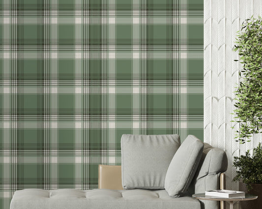 A modern living space featuring a prominent olive green and white plaid wallpaper accent wall. In front of the wall sits a sleek grey chaise lounge with matching plush pillows, accompanied by a small, round metal side table. To the left, a large window is dressed with sheer white curtains and long, sage green drapes over herringbone wood flooring. To the right, a textured white feature wall provides contrast, with a large potted green plant in a dark ceramic planter standing in the corner.