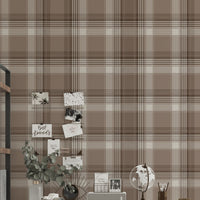 A styled home office area featuring a prominent faded mocha brown and white plaid wallpaper. In the center, a white writing desk is paired with a matching white chair draped with a chunky beige knit blanket. The desk is organized with a small globe, a desk organizer, a camera, and a vase with eucalyptus branches. To the left stands a tall industrial-style wooden bookshelf with a black metal frame, holding books and decor. To the right, a silver metal task lamp rests on a small black side table near an open 