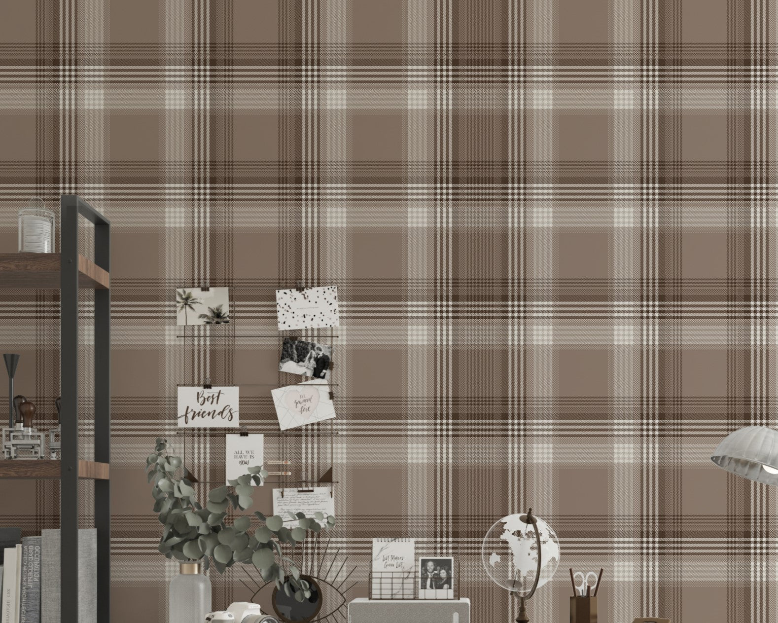 A styled home office area featuring a prominent faded mocha brown and white plaid wallpaper. In the center, a white writing desk is paired with a matching white chair draped with a chunky beige knit blanket. The desk is organized with a small globe, a desk organizer, a camera, and a vase with eucalyptus branches. To the left stands a tall industrial-style wooden bookshelf with a black metal frame, holding books and decor. To the right, a silver metal task lamp rests on a small black side table near an open 