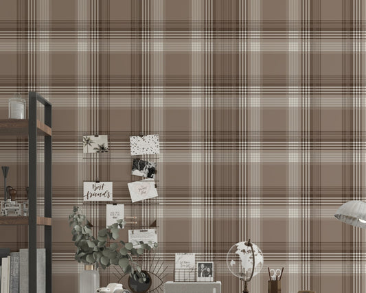 A styled home office area featuring a prominent faded mocha brown and white plaid wallpaper. In the center, a white writing desk is paired with a matching white chair draped with a chunky beige knit blanket. The desk is organized with a small globe, a desk organizer, a camera, and a vase with eucalyptus branches. To the left stands a tall industrial-style wooden bookshelf with a black metal frame, holding books and decor. To the right, a silver metal task lamp rests on a small black side table near an open 