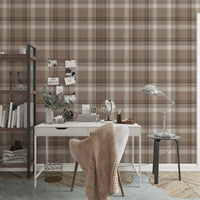 A styled home office area featuring a prominent faded mocha brown and white plaid wallpaper. In the center, a white writing desk is paired with a matching white chair draped with a chunky beige knit blanket. The desk is organized with a small globe, a desk organizer, a camera, and a vase with eucalyptus branches. To the left stands a tall industrial-style wooden bookshelf with a black metal frame, holding books and decor. To the right, a silver metal task lamp rests on a small black side table near an open 