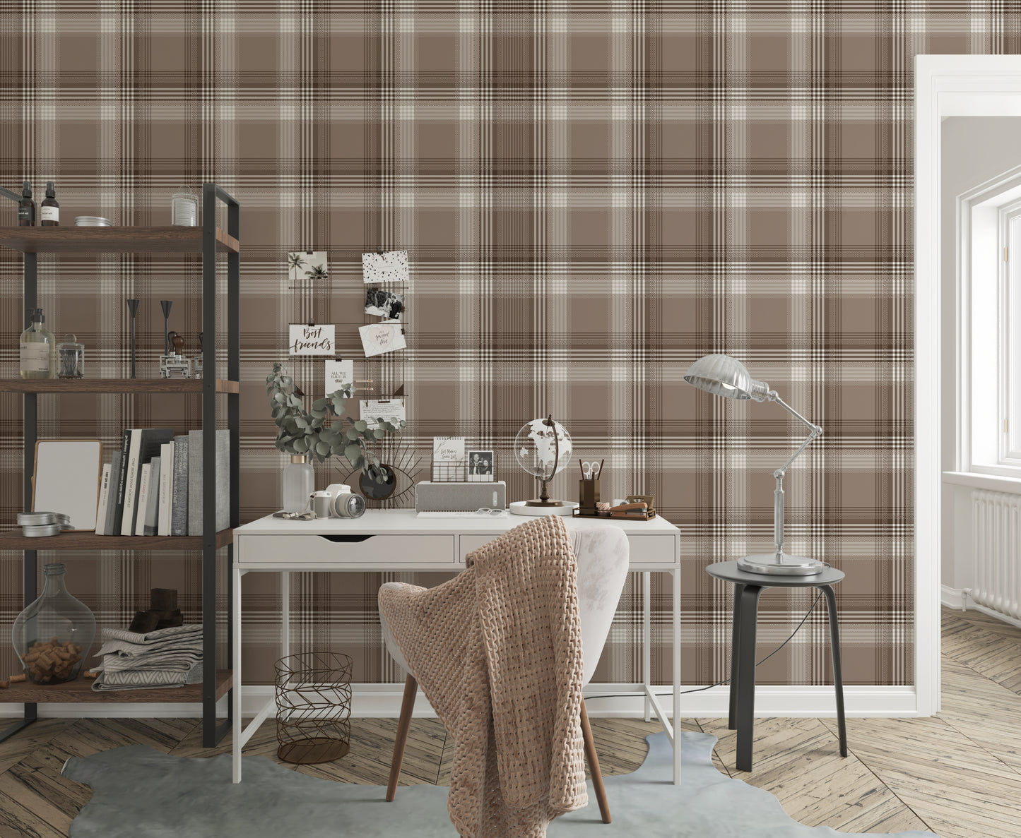 A styled home office area featuring a prominent faded mocha brown and white plaid wallpaper. In the center, a white writing desk is paired with a matching white chair draped with a chunky beige knit blanket. The desk is organized with a small globe, a desk organizer, a camera, and a vase with eucalyptus branches. To the left stands a tall industrial-style wooden bookshelf with a black metal frame, holding books and decor. To the right, a silver metal task lamp rests on a small black side table near an open 