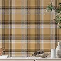 A closeup of a curated tabletop setting featuring a warm honey-gold plaid wallpaper with brown and cream accents. On the light-colored console surface, an assortment of earth-toned decor is arranged, including a small terracotta footed bowl on a white book to the left. To the right sits a collection of ribbed ceramic vases in white and cream, a shallow decorative bowl containing a sprig of dried pampas grass, and a tall, dark glass vase holding green leafy branches.