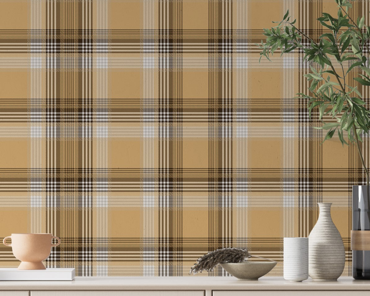 A closeup of a curated tabletop setting featuring a warm honey-gold plaid wallpaper with brown and cream accents. On the light-colored console surface, an assortment of earth-toned decor is arranged, including a small terracotta footed bowl on a white book to the left. To the right sits a collection of ribbed ceramic vases in white and cream, a shallow decorative bowl containing a sprig of dried pampas grass, and a tall, dark glass vase holding green leafy branches.