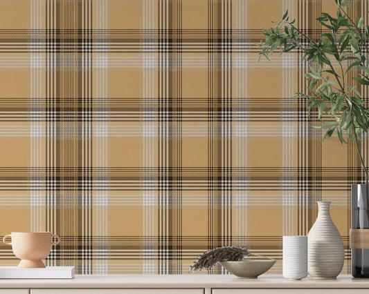 A closeup of a curated tabletop setting featuring a warm honey-gold plaid wallpaper with brown and cream accents. On the light-colored console surface, an assortment of earth-toned decor is arranged, including a small terracotta footed bowl on a white book to the left. To the right sits a collection of ribbed ceramic vases in white and cream, a shallow decorative bowl containing a sprig of dried pampas grass, and a tall, dark glass vase holding green leafy branches.