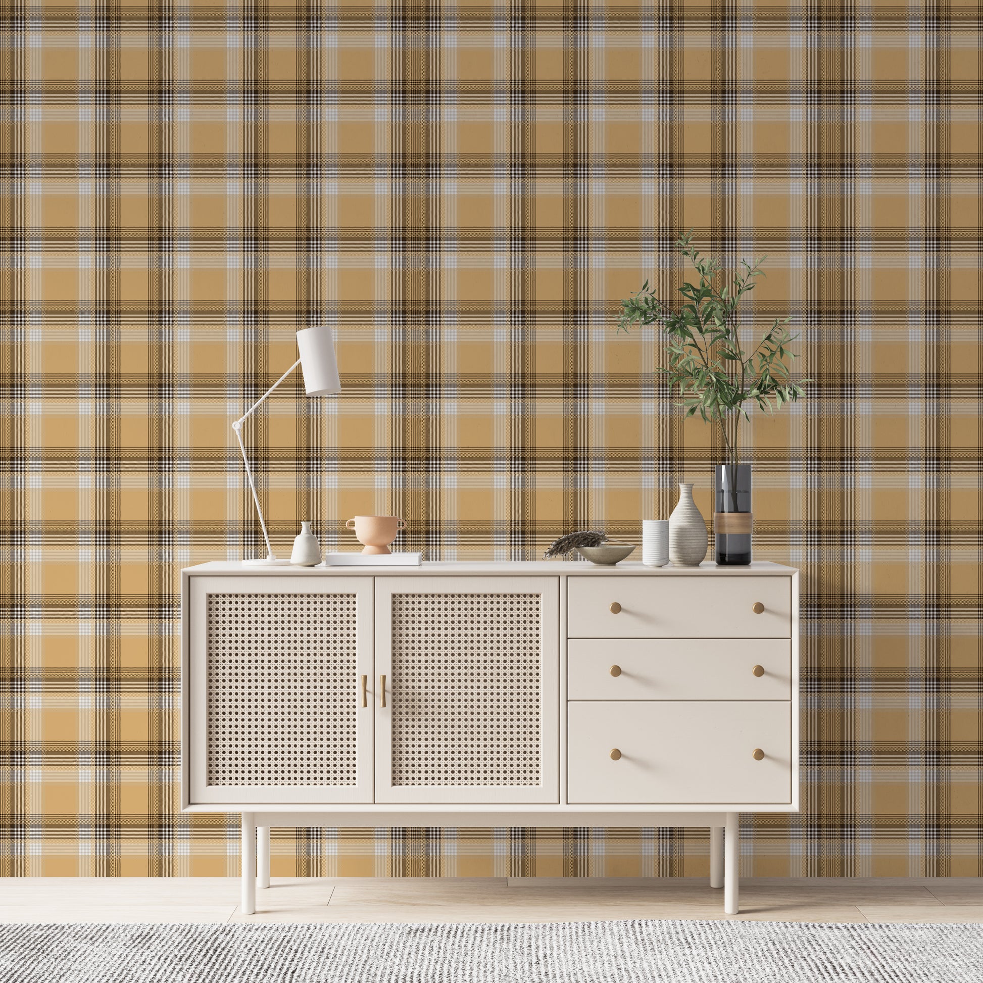 A closeup of a curated tabletop setting featuring a warm honey-gold plaid wallpaper with brown and cream accents. On the light-colored console surface, an assortment of earth-toned decor is arranged, including a small terracotta footed bowl on a white book to the left. To the right sits a collection of ribbed ceramic vases in white and cream, a shallow decorative bowl containing a sprig of dried pampas grass, and a tall, dark glass vase holding green leafy branches.