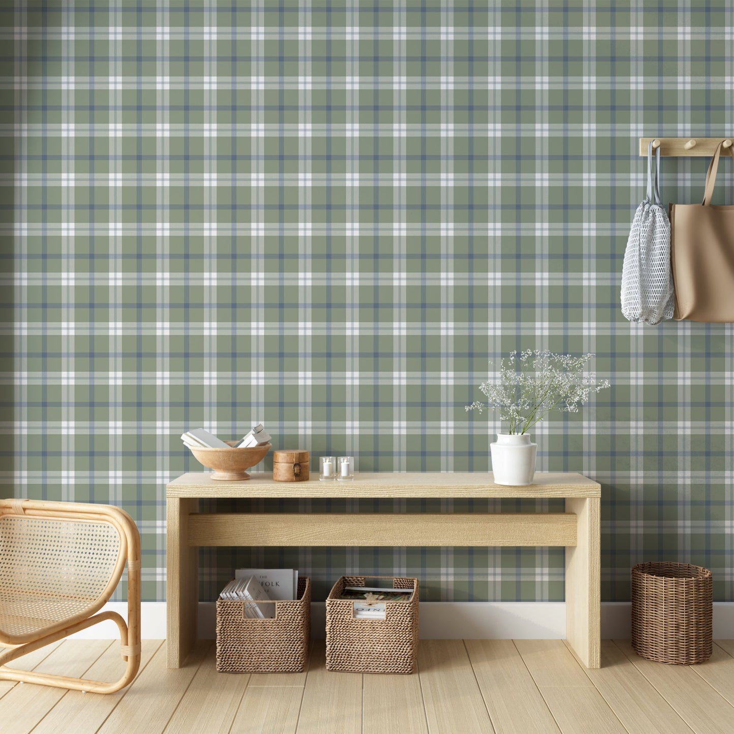 A cozy, rustic room vignette featuring a wall covered in a classic plaid wallpaper pattern of muted stone green, navy blue, and white. In the foreground, a light-colored wooden console table displays a wooden bowl filled with books, a small round wooden box with a latch, and two small votive candles in clear glass holders. The corner of a woven rattan chair is visible on the bottom left.