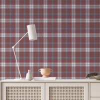A cozy and styled room interior featuring a wall covered in a classic plaid wallpaper with a muted brick red background, crisscrossed with blue, grey, and white stripes. In the foreground, a cream-colored wooden sideboard with woven rattan mesh cabinet doors displays a modern white desk lamp, a small beige ceramic vase, a footed tan bowl sitting on a book, and a small decorative dish with dried botanicals.