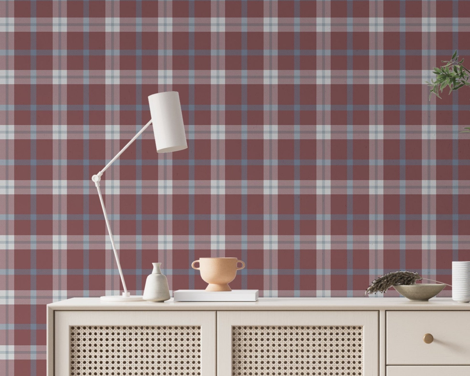 A cozy and styled room interior featuring a wall covered in a classic plaid wallpaper with a muted brick red background, crisscrossed with blue, grey, and white stripes. In the foreground, a cream-colored wooden sideboard with woven rattan mesh cabinet doors displays a modern white desk lamp, a small beige ceramic vase, a footed tan bowl sitting on a book, and a small decorative dish with dried botanicals.
