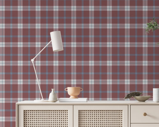 A cozy and styled room interior featuring a wall covered in a classic plaid wallpaper with a muted brick red background, crisscrossed with blue, grey, and white stripes. In the foreground, a cream-colored wooden sideboard with woven rattan mesh cabinet doors displays a modern white desk lamp, a small beige ceramic vase, a footed tan bowl sitting on a book, and a small decorative dish with dried botanicals.