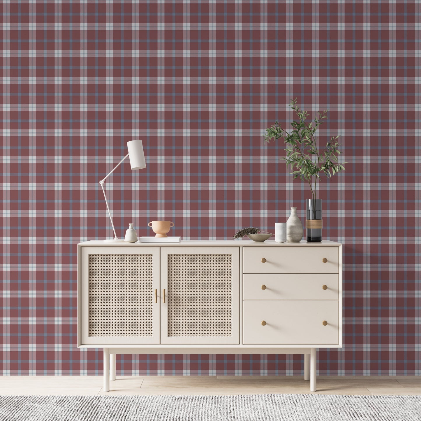 A cozy and styled room interior featuring a wall covered in a classic plaid wallpaper with a muted brick red background, crisscrossed with blue, grey, and white stripes. In the foreground, a cream-colored wooden sideboard with woven rattan mesh cabinet doors displays a modern white desk lamp, a small beige ceramic vase, a footed tan bowl sitting on a book, and a small decorative dish with dried botanicals.