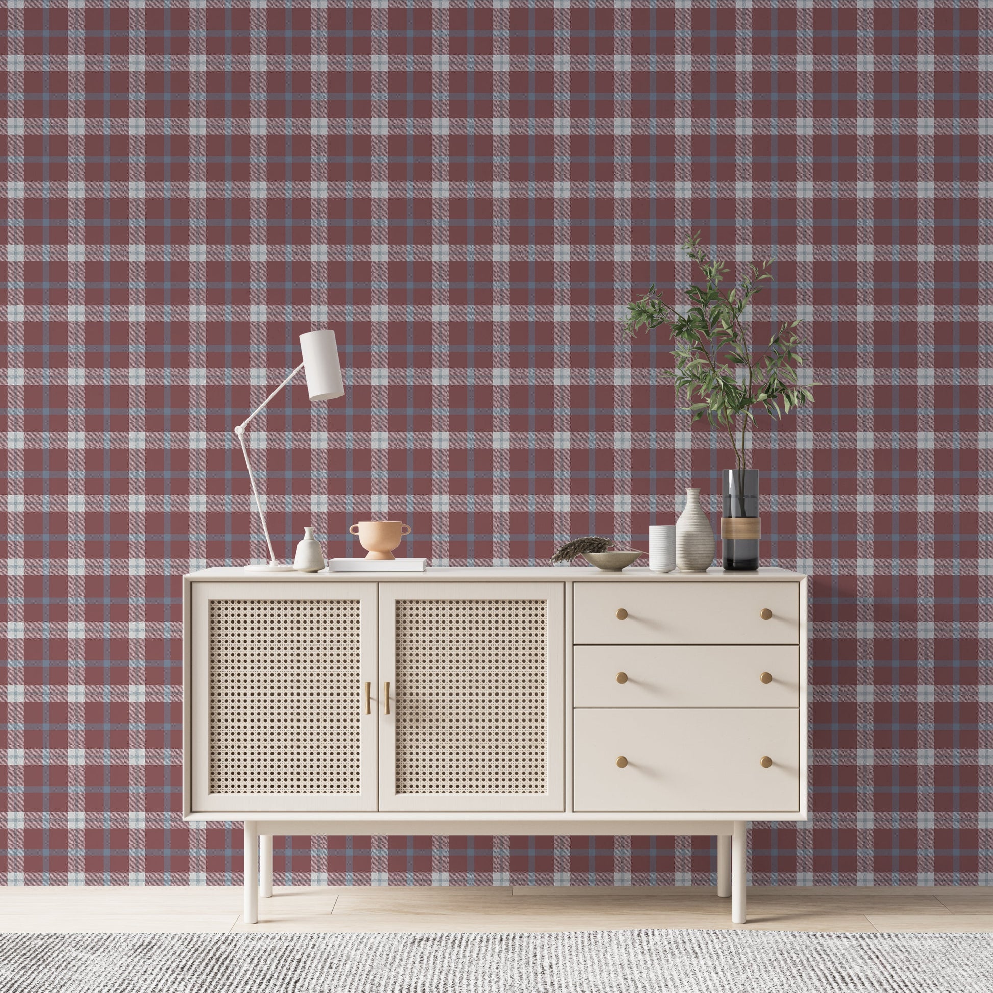 A cozy and styled room interior featuring a wall covered in a classic plaid wallpaper with a muted brick red background, crisscrossed with blue, grey, and white stripes. In the foreground, a cream-colored wooden sideboard with woven rattan mesh cabinet doors displays a modern white desk lamp, a small beige ceramic vase, a footed tan bowl sitting on a book, and a small decorative dish with dried botanicals.