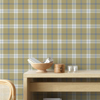 A rustic room vignette featuring a wall covered in a classic plaid wallpaper pattern with an olive tan background crisscrossed by muted blue and white lines. In the foreground, a light-colored wooden console table displays a wooden bowl filled with books, a small round wooden box with a latch, and two small votive candles in clear glass holders. The corner of a woven rattan chair is visible on the bottom left.