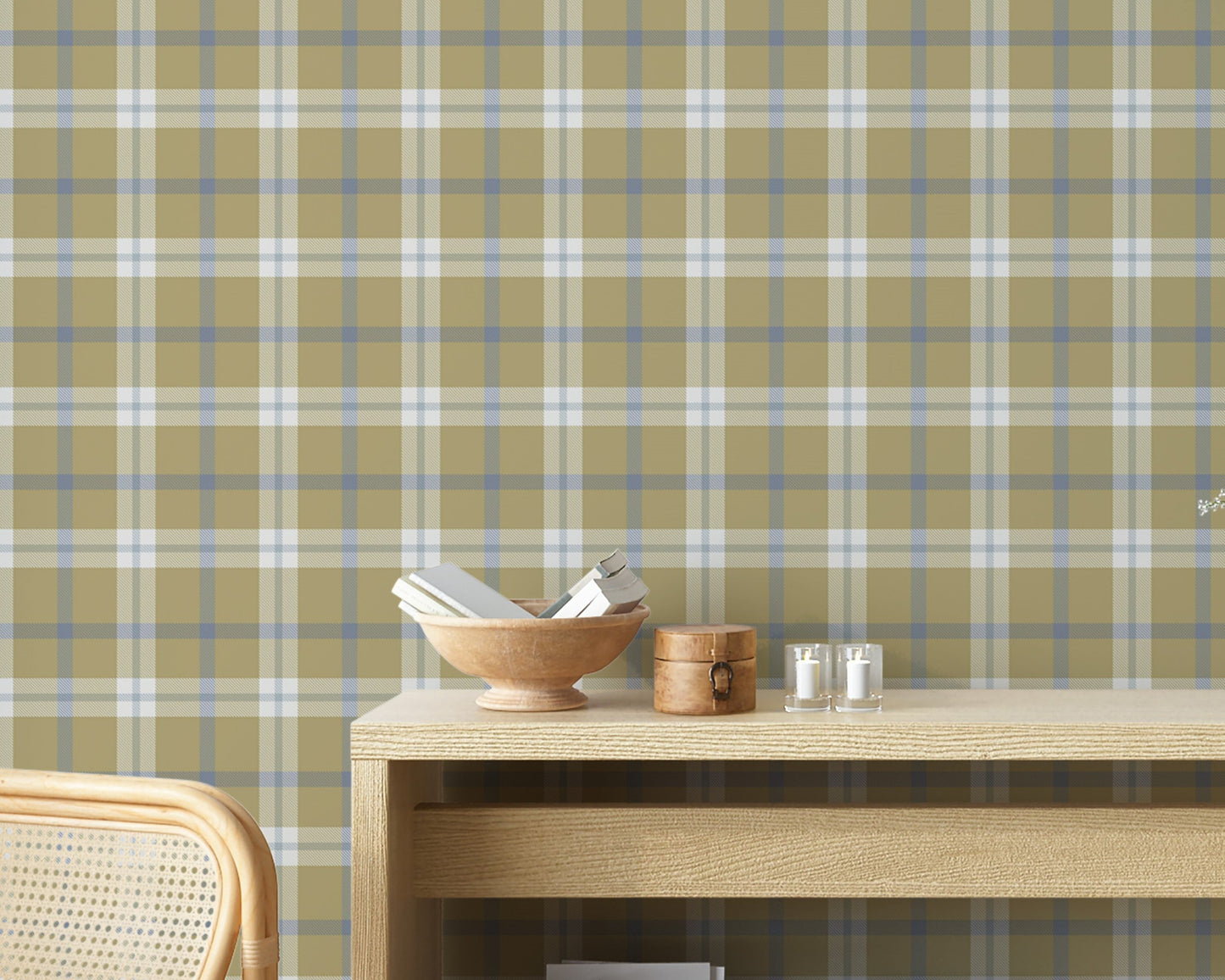 A rustic room vignette featuring a wall covered in a classic plaid wallpaper pattern with an olive tan background crisscrossed by muted blue and white lines. In the foreground, a light-colored wooden console table displays a wooden bowl filled with books, a small round wooden box with a latch, and two small votive candles in clear glass holders. The corner of a woven rattan chair is visible on the bottom left.