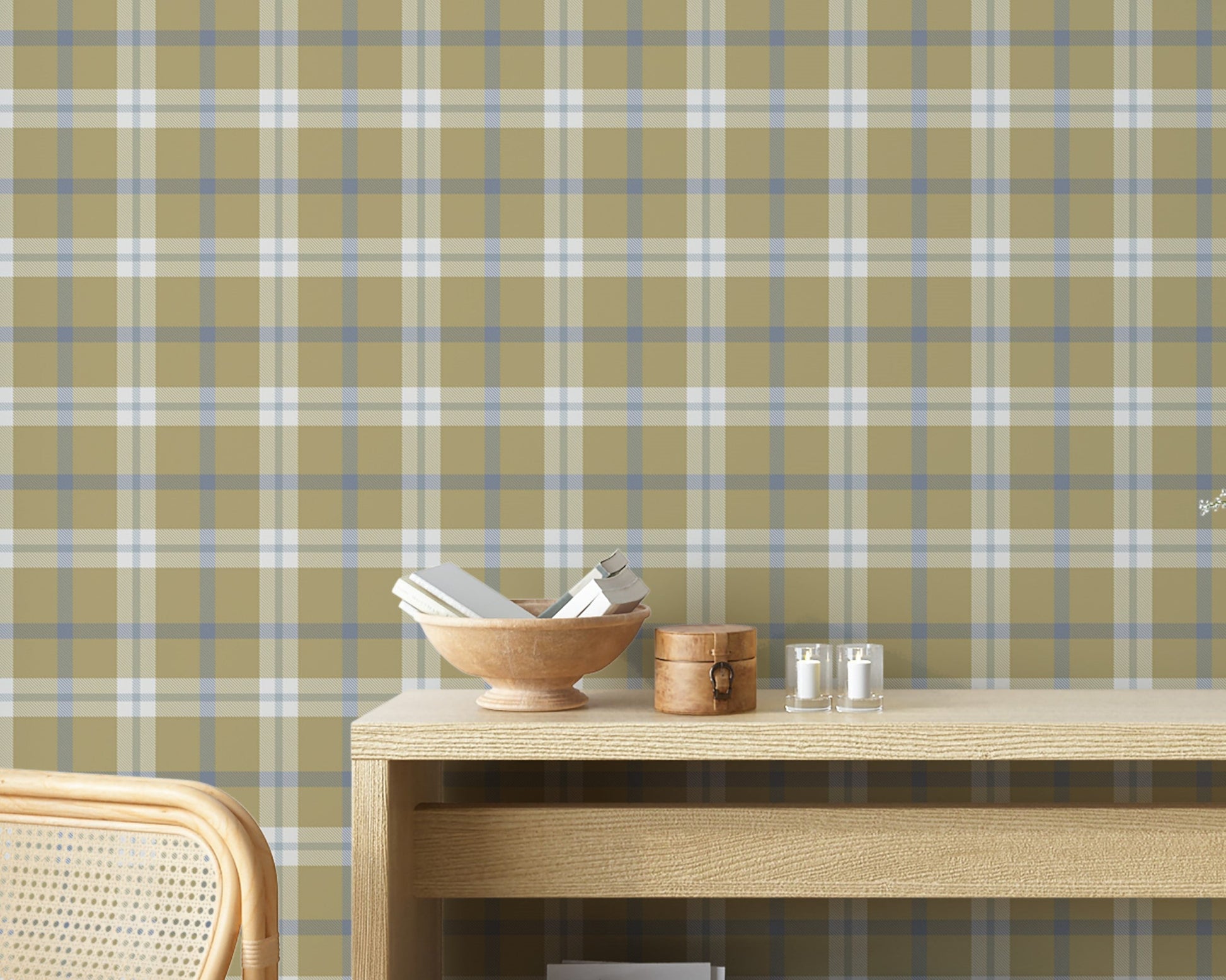 A rustic room vignette featuring a wall covered in a classic plaid wallpaper pattern with an olive tan background crisscrossed by muted blue and white lines. In the foreground, a light-colored wooden console table displays a wooden bowl filled with books, a small round wooden box with a latch, and two small votive candles in clear glass holders. The corner of a woven rattan chair is visible on the bottom left.