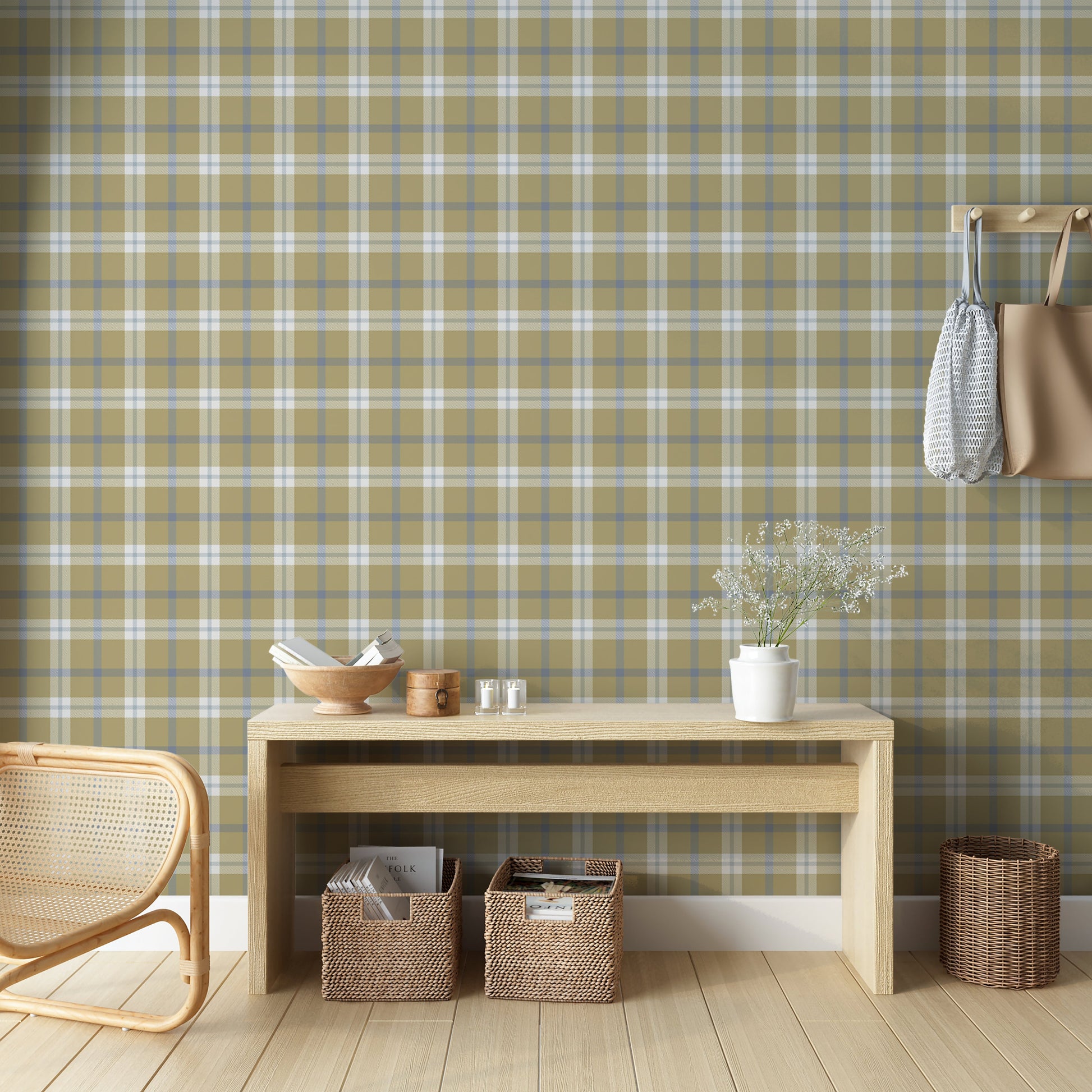 A rustic room vignette featuring a wall covered in a classic plaid wallpaper pattern with an olive tan background crisscrossed by muted blue and white lines. In the foreground, a light-colored wooden console table displays a wooden bowl filled with books, a small round wooden box with a latch, and two small votive candles in clear glass holders. The corner of a woven rattan chair is visible on the bottom left.