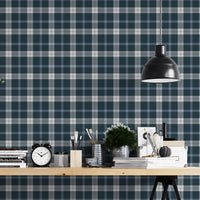 A modern studio or workspace setting featuring a wall covered in a classic plaid wallpaper pattern with a midnight steel blue background, crisscrossed by white and grey stripes. In the foreground, a light-colored wooden desk is arranged with various items, including a stack of books, a black vintage-style alarm clock, paintbrushes in holders, a small potted green plant, and storage boxes. A matte black dome pendant light hangs above the right side of the desk, illuminating the workspace.