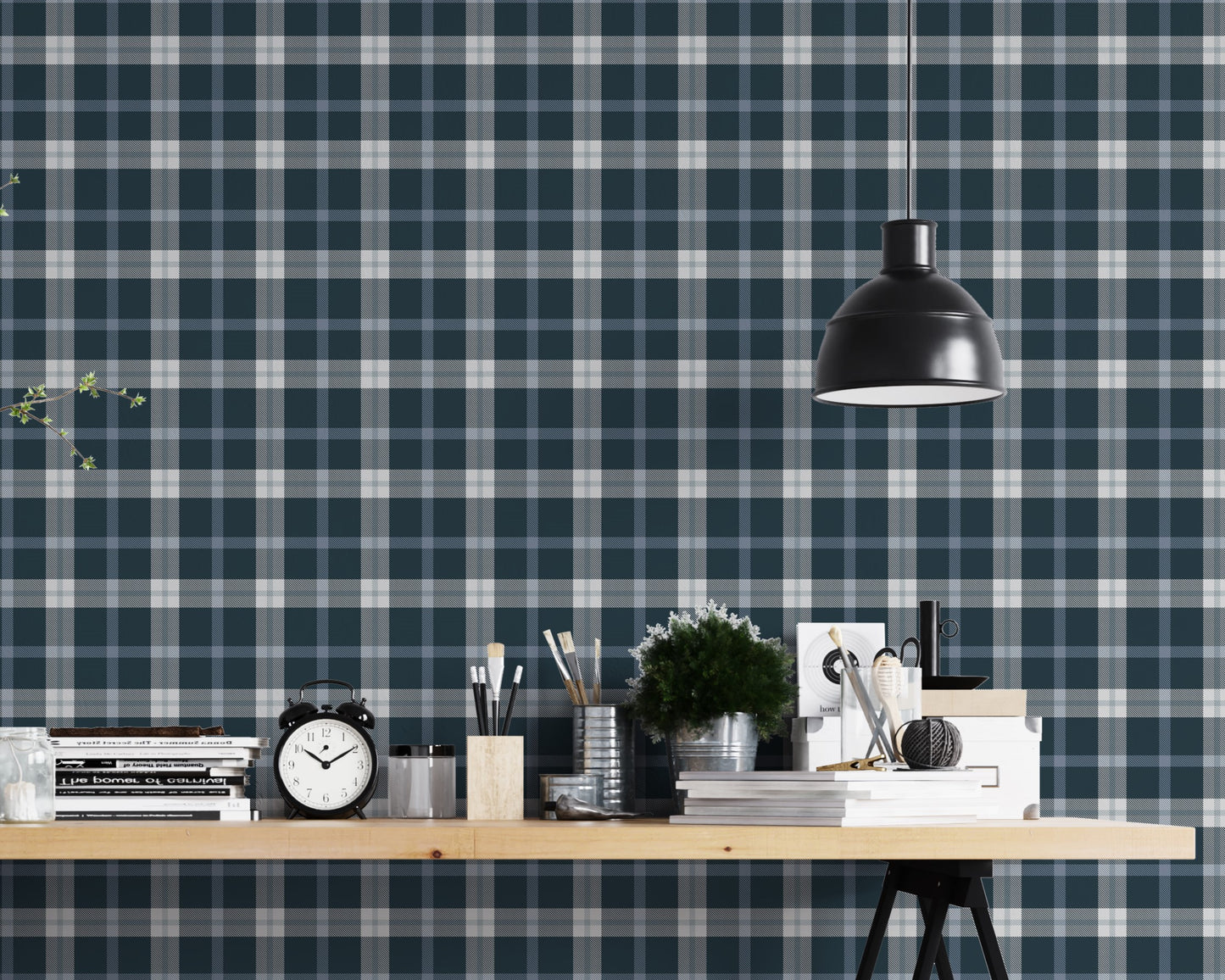 A modern studio or workspace setting featuring a wall covered in a classic plaid wallpaper pattern with a midnight steel blue background, crisscrossed by white and grey stripes. In the foreground, a light-colored wooden desk is arranged with various items, including a stack of books, a black vintage-style alarm clock, paintbrushes in holders, a small potted green plant, and storage boxes. A matte black dome pendant light hangs above the right side of the desk, illuminating the workspace.