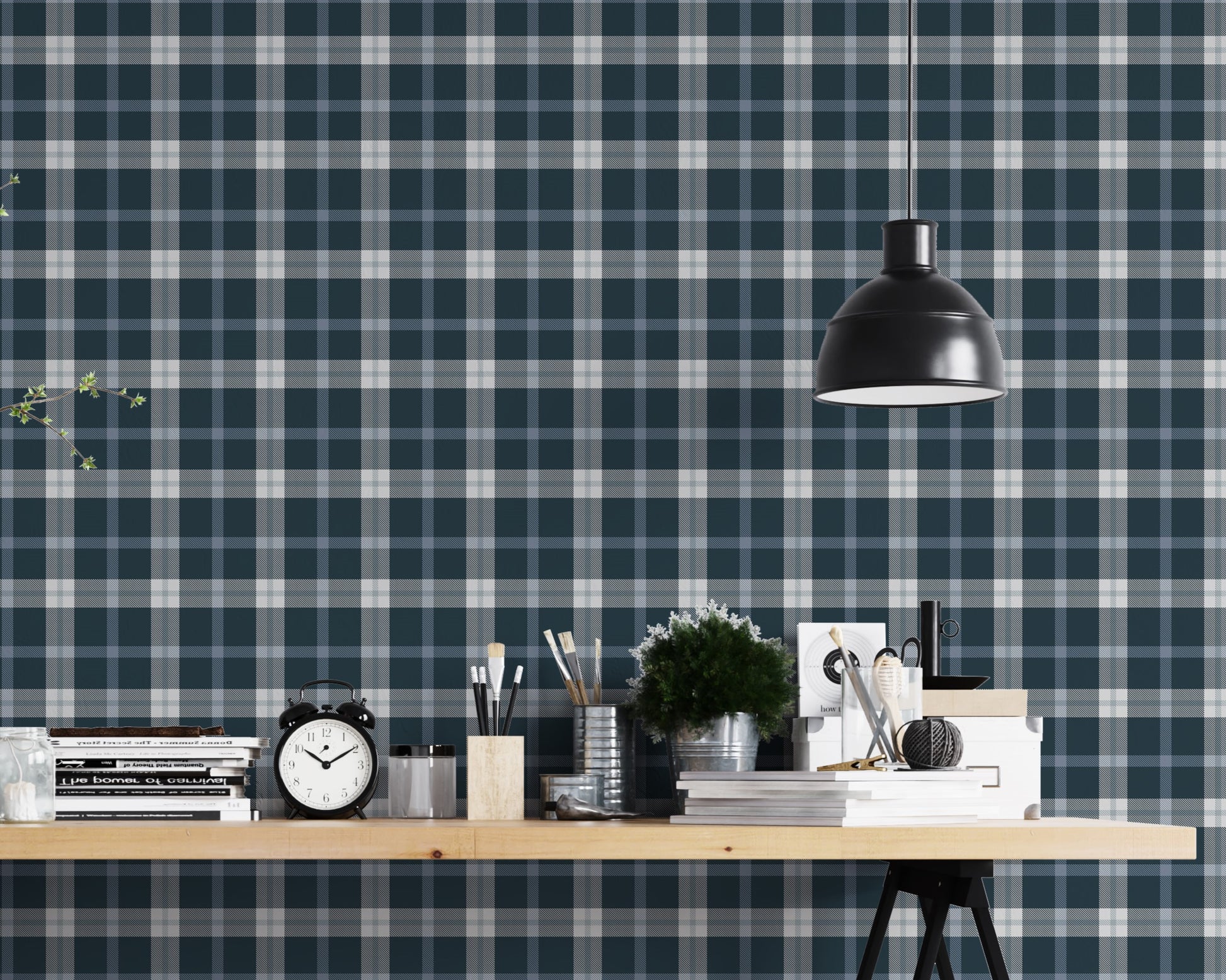 A modern studio or workspace setting featuring a wall covered in a classic plaid wallpaper pattern with a midnight steel blue background, crisscrossed by white and grey stripes. In the foreground, a light-colored wooden desk is arranged with various items, including a stack of books, a black vintage-style alarm clock, paintbrushes in holders, a small potted green plant, and storage boxes. A matte black dome pendant light hangs above the right side of the desk, illuminating the workspace.