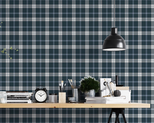 A modern studio or workspace setting featuring a wall covered in a classic plaid wallpaper pattern with a midnight steel blue background, crisscrossed by white and grey stripes. In the foreground, a light-colored wooden desk is arranged with various items, including a stack of books, a black vintage-style alarm clock, paintbrushes in holders, a small potted green plant, and storage boxes. A matte black dome pendant light hangs above the right side of the desk, illuminating the workspace.