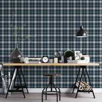 A modern studio or workspace setting featuring a wall covered in a classic plaid wallpaper pattern with a midnight steel blue background, crisscrossed by white and grey stripes. In the foreground, a light-colored wooden desk is arranged with various items, including a stack of books, a black vintage-style alarm clock, paintbrushes in holders, a small potted green plant, and storage boxes. A matte black dome pendant light hangs above the right side of the desk, illuminating the workspace.