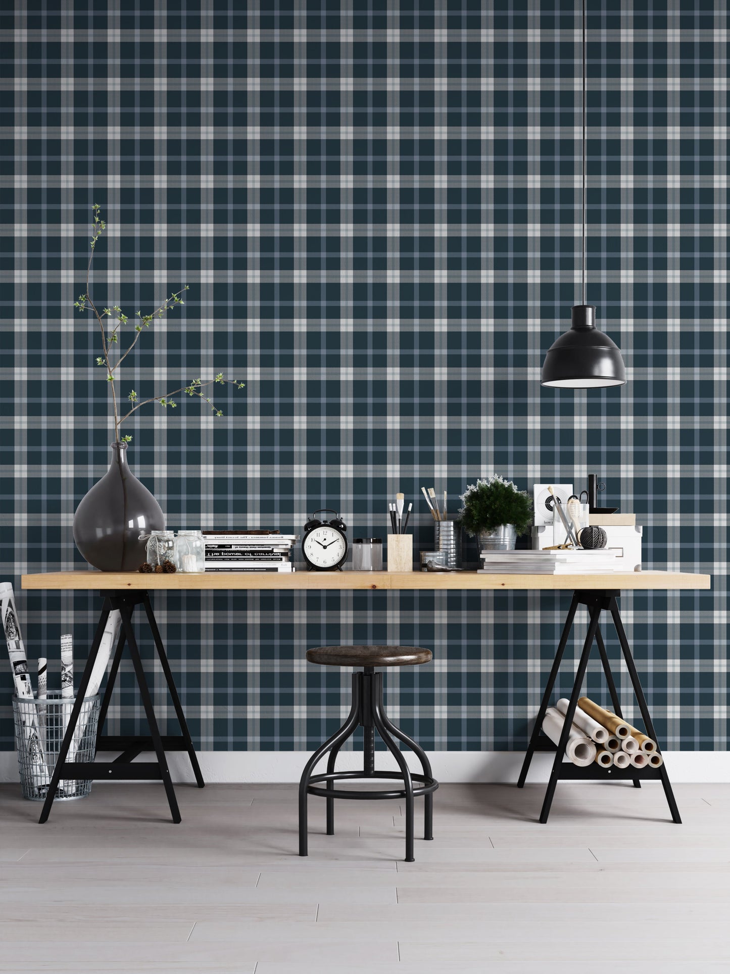 A modern studio or workspace setting featuring a wall covered in a classic plaid wallpaper pattern with a midnight steel blue background, crisscrossed by white and grey stripes. In the foreground, a light-colored wooden desk is arranged with various items, including a stack of books, a black vintage-style alarm clock, paintbrushes in holders, a small potted green plant, and storage boxes. A matte black dome pendant light hangs above the right side of the desk, illuminating the workspace.