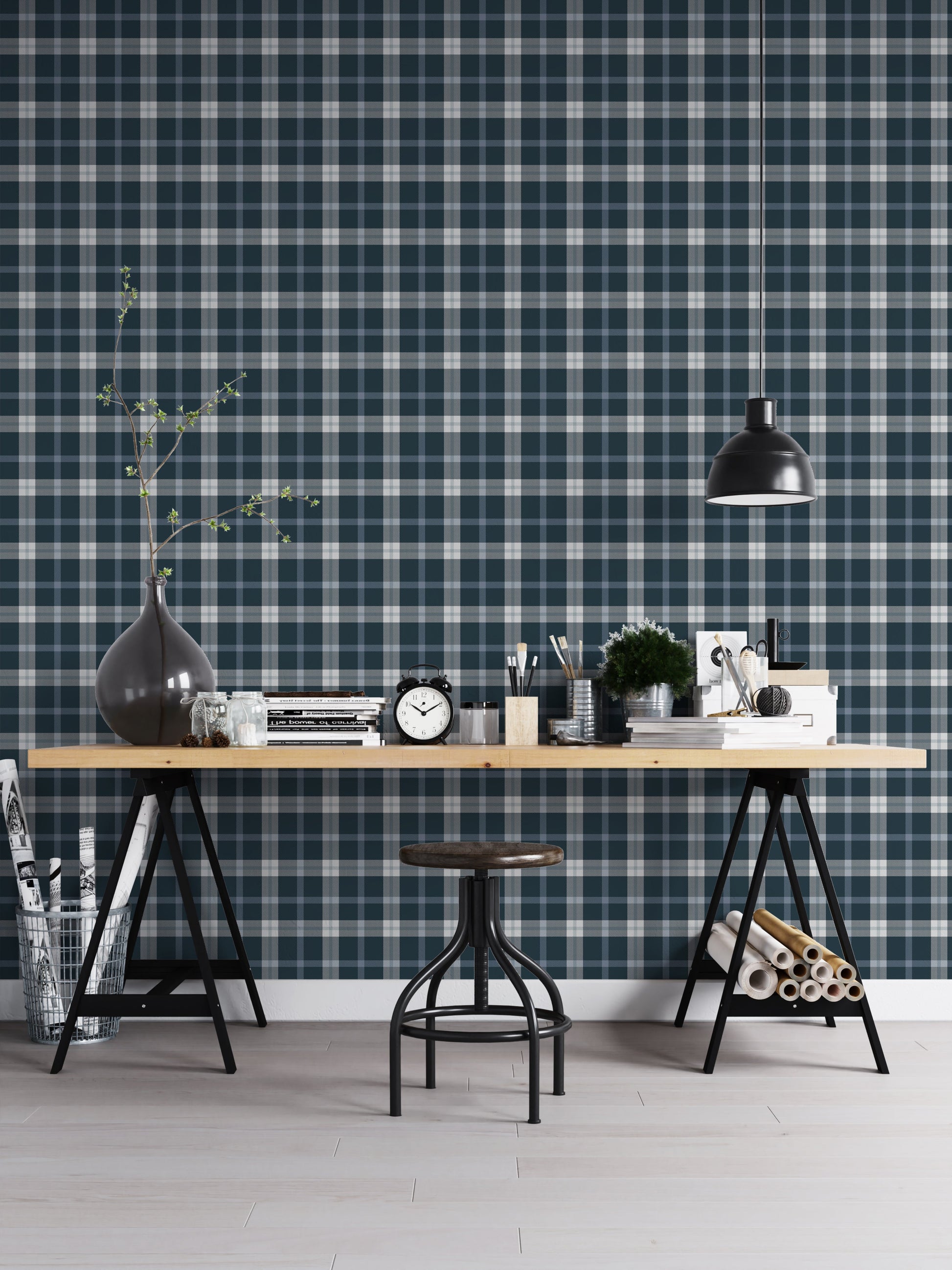 A modern studio or workspace setting featuring a wall covered in a classic plaid wallpaper pattern with a midnight steel blue background, crisscrossed by white and grey stripes. In the foreground, a light-colored wooden desk is arranged with various items, including a stack of books, a black vintage-style alarm clock, paintbrushes in holders, a small potted green plant, and storage boxes. A matte black dome pendant light hangs above the right side of the desk, illuminating the workspace.