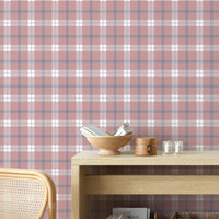 A cozy and styled room interior featuring a wall covered in a classic plaid wallpaper with a muted brick red background, crisscrossed with blue, grey, and white stripes. In the foreground, a cream-colored wooden sideboard with woven rattan mesh cabinet doors displays a modern white desk lamp, a small beige ceramic vase, a footed tan bowl sitting on a book, and a small decorative dish with dried botanicals.