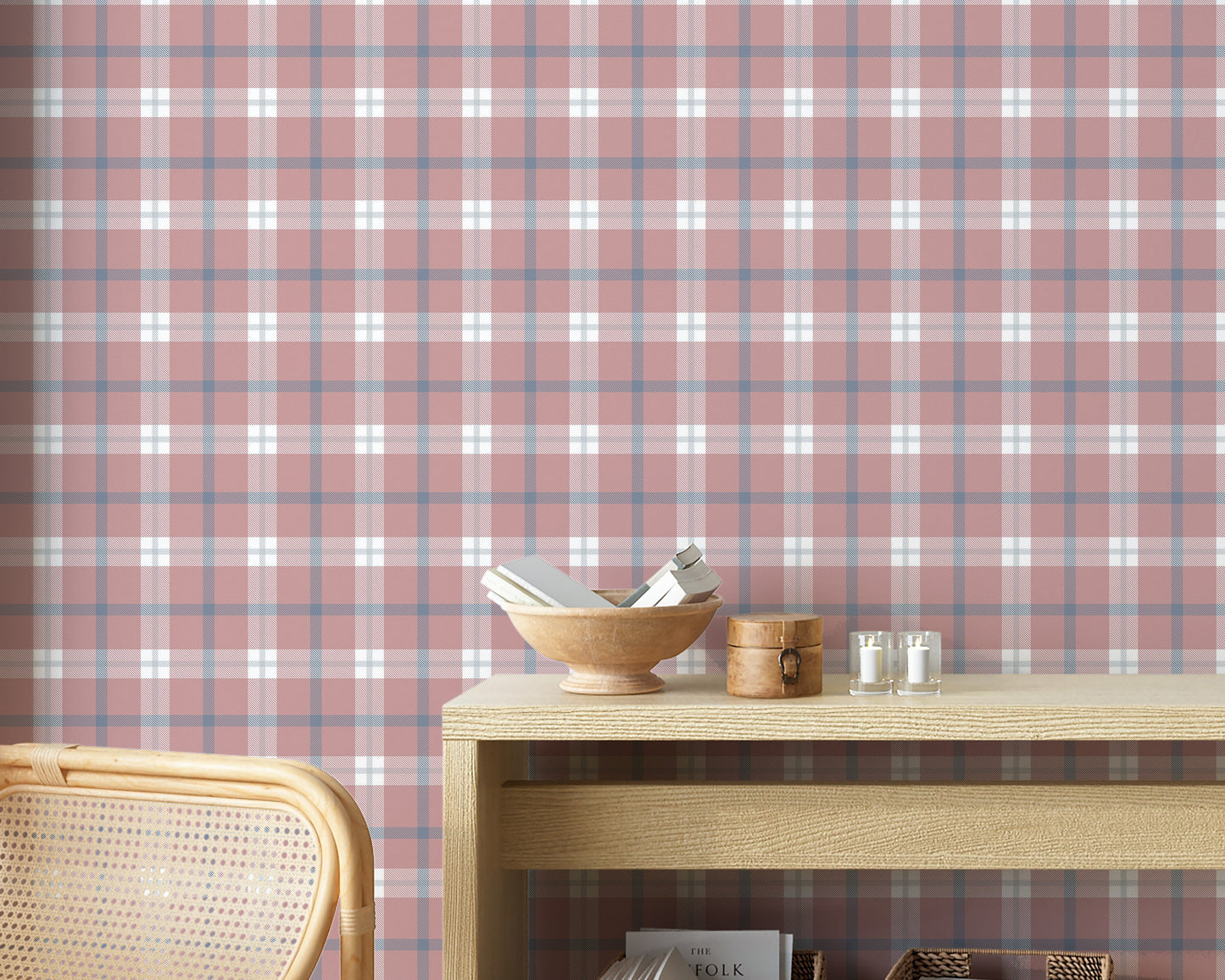 A cozy and styled room interior featuring a wall covered in a classic plaid wallpaper with a muted brick red background, crisscrossed with blue, grey, and white stripes. In the foreground, a cream-colored wooden sideboard with woven rattan mesh cabinet doors displays a modern white desk lamp, a small beige ceramic vase, a footed tan bowl sitting on a book, and a small decorative dish with dried botanicals.