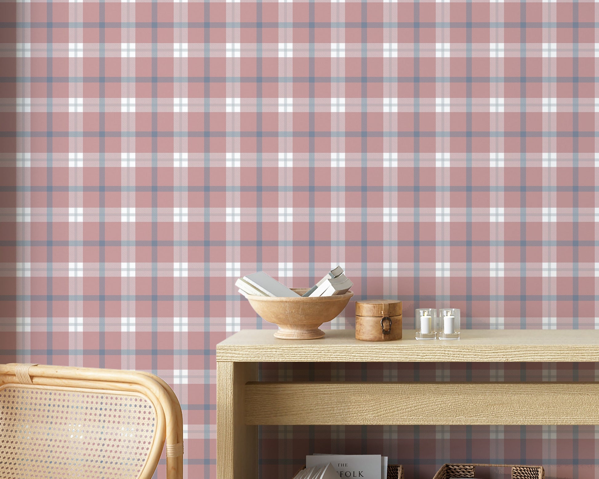 A cozy and styled room interior featuring a wall covered in a classic plaid wallpaper with a muted brick red background, crisscrossed with blue, grey, and white stripes. In the foreground, a cream-colored wooden sideboard with woven rattan mesh cabinet doors displays a modern white desk lamp, a small beige ceramic vase, a footed tan bowl sitting on a book, and a small decorative dish with dried botanicals.