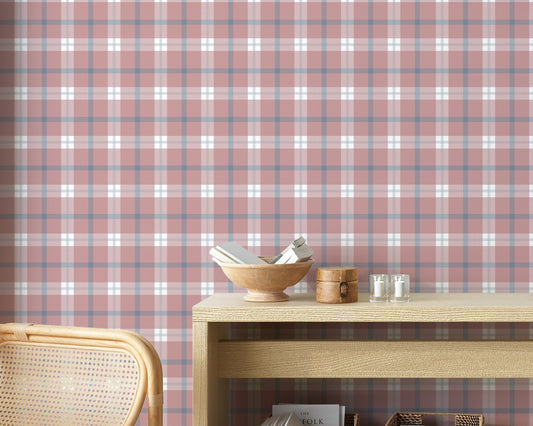 A cozy and styled room interior featuring a wall covered in a classic plaid wallpaper with a muted brick red background, crisscrossed with blue, grey, and white stripes. In the foreground, a cream-colored wooden sideboard with woven rattan mesh cabinet doors displays a modern white desk lamp, a small beige ceramic vase, a footed tan bowl sitting on a book, and a small decorative dish with dried botanicals.