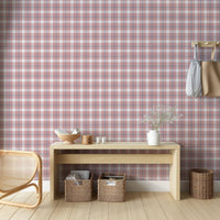 A cozy and styled room interior featuring a wall covered in a classic plaid wallpaper with a muted brick red background, crisscrossed with blue, grey, and white stripes. In the foreground, a cream-colored wooden sideboard with woven rattan mesh cabinet doors displays a modern white desk lamp, a small beige ceramic vase, a footed tan bowl sitting on a book, and a small decorative dish with dried botanicals.