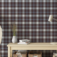 A neatly styled console table setting set against a wall with a classic plaid wallpaper pattern in a smoky cocoa brown background, layered with light blue and white stripes. On the light-colored wooden table sits a white ribbed vase with dried grass stalks, a small stack of books, and a modern cream-colored desk lamp on the right. Below the table, woven storage baskets filled with books are visible on a lower shelf, and a partial view of large woven leaf-shaped decor stands to the left.