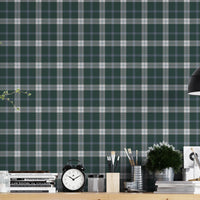 A modern workspace or studio setting featuring a wall covered in a classic plaid wallpaper pattern with a deep juniper green background, crisscrossed by white and muted grey stripes. In the foreground, a light-colored wooden desk is organized with various items, including stacks of books, a black vintage-style twin-bell alarm clock, small jars, paintbrushes in containers, and a small potted green plant. A matte black dome pendant light hangs over the right side of the desk, illuminating the workspace.