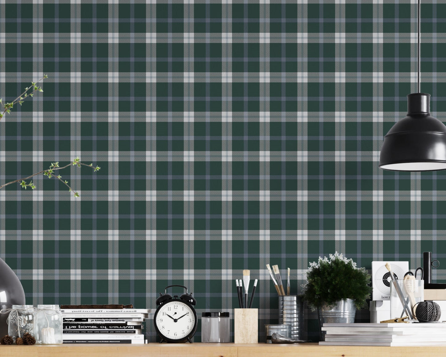 A modern workspace or studio setting featuring a wall covered in a classic plaid wallpaper pattern with a deep juniper green background, crisscrossed by white and muted grey stripes. In the foreground, a light-colored wooden desk is organized with various items, including stacks of books, a black vintage-style twin-bell alarm clock, small jars, paintbrushes in containers, and a small potted green plant. A matte black dome pendant light hangs over the right side of the desk, illuminating the workspace.
