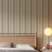 A warm, inviting bedroom scene showcasing a light wood paneled headboard against a wall covered in vertical dusty beige, cream, and muted olive green striped wallpaper. The bed is dressed with crisp white sheets, a brown duvet, and a dusty pink pillow. To the right, a wooden nightstand holds a small candle, a small reddish-brown glass vase, and a textured ceramic vase, all illuminated by the soft, warm glow of a round orb wall sconce.