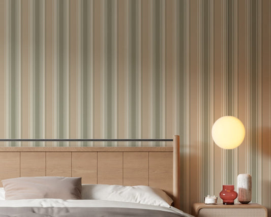 A warm, inviting bedroom scene showcasing a light wood paneled headboard against a wall covered in vertical dusty beige, cream, and muted olive green striped wallpaper. The bed is dressed with crisp white sheets, a brown duvet, and a dusty pink pillow. To the right, a wooden nightstand holds a small candle, a small reddish-brown glass vase, and a textured ceramic vase, all illuminated by the soft, warm glow of a round orb wall sconce.