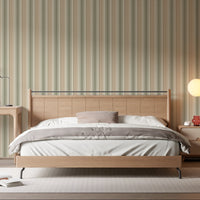 A warm, inviting bedroom scene showcasing a light wood paneled headboard against a wall covered in vertical dusty beige, cream, and muted olive green striped wallpaper. The bed is dressed with crisp white sheets, a brown duvet, and a dusty pink pillow. To the right, a wooden nightstand holds a small candle, a small reddish-brown glass vase, and a textured ceramic vase, all illuminated by the soft, warm glow of a round orb wall sconce.
