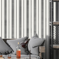 A modern interior scene with a gray cushioned sofa set against a white wall patterned with thin vertical black stripes. A wooden side table and black desk lamp sit beside bottles and glasses on a coffee table, while an industrial-style shelving unit with metal mesh panels stands on the right.