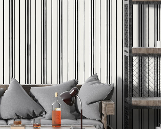 A modern interior scene with a gray cushioned sofa set against a white wall patterned with thin vertical black stripes. A wooden side table and black desk lamp sit beside bottles and glasses on a coffee table, while an industrial-style shelving unit with metal mesh panels stands on the right.