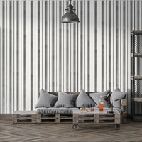A modern interior scene with a gray cushioned sofa set against a white wall patterned with thin vertical black stripes. A wooden side table and black desk lamp sit beside bottles and glasses on a coffee table, while an industrial-style shelving unit with metal mesh panels stands on the right.