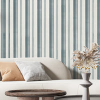 A contemporary living room with a curved white sofa accented by beige and gray throw pillows. Behind the sofa is a white wall with thin vertical teal stripes, while a white vase filled with dried pampas grass and yellow berry stems sits on a table in the foreground.