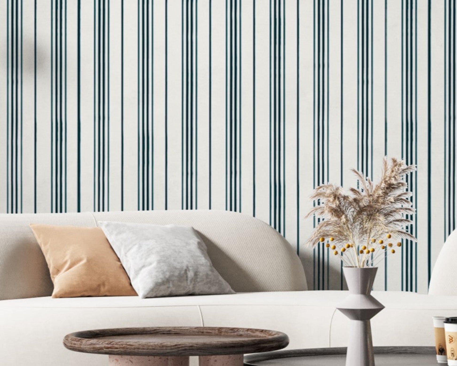 A contemporary living room with a curved white sofa accented by beige and gray throw pillows. Behind the sofa is a white wall with thin vertical teal stripes, while a white vase filled with dried pampas grass and yellow berry stems sits on a table in the foreground.
