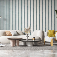 A contemporary living room with a curved white sofa accented by beige and gray throw pillows. Behind the sofa is a white wall with thin vertical teal stripes, while a white vase filled with dried pampas grass and yellow berry stems sits on a table in the foreground.