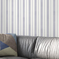 A gray leather sofa with a geometric-patterned throw pillow sits against a white wall decorated with thin vertical lavender-gray stripes. The minimalist interior features soft textures and a modern, neutral-toned design.