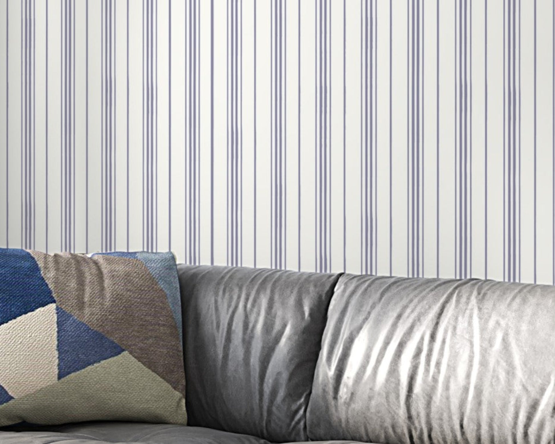 A gray leather sofa with a geometric-patterned throw pillow sits against a white wall decorated with thin vertical lavender-gray stripes. The minimalist interior features soft textures and a modern, neutral-toned design.