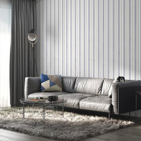 A gray leather sofa with a geometric-patterned throw pillow sits against a white wall decorated with thin vertical lavender-gray stripes. The minimalist interior features soft textures and a modern, neutral-toned design.