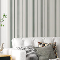 An airy, modern living room featuring a white tufted sofa against a wall with vertical grey and olive green stripes. The sofa is adorned with a mix of plaid, checkered, and solid olive-colored throw pillows, along with a draped plaid blanket. It sits on a dark grey patterned area rug over light-colored hardwood floors. To the left is a small black and light wood side table holding a vase and a bird figurine. On the right, a large, black metal-framed glass sliding partition separates the space from another 
