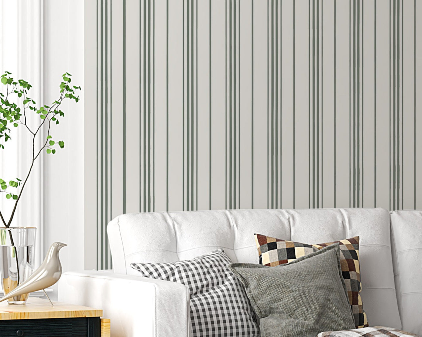 An airy, modern living room featuring a white tufted sofa against a wall with vertical grey and olive green stripes. The sofa is adorned with a mix of plaid, checkered, and solid olive-colored throw pillows, along with a draped plaid blanket. It sits on a dark grey patterned area rug over light-colored hardwood floors. To the left is a small black and light wood side table holding a vase and a bird figurine. On the right, a large, black metal-framed glass sliding partition separates the space from another 