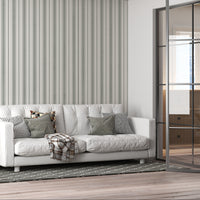 An airy, modern living room featuring a white tufted sofa against a wall with vertical grey and olive green stripes. The sofa is adorned with a mix of plaid, checkered, and solid olive-colored throw pillows, along with a draped plaid blanket. It sits on a dark grey patterned area rug over light-colored hardwood floors. To the left is a small black and light wood side table holding a vase and a bird figurine. On the right, a large, black metal-framed glass sliding partition separates the space from another 